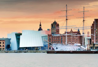 Moderne Museumsarchitektur trifft auf hanseatisches Erbe: Das Ozeaneum am Stralsunder Hafen verschmilzt mit historischen Backsteinbauten und Segelschiffen zu einer einzigartigen Silhouette. Im Abendlicht leuchtet die UNESCO-Welterbestadt in warmen Farbt&ouml;nen. Hier l&auml;sst sich maritime Geschichte hautnah erleben &ndash; vom Museumsdeck bis zur Hafenpromenade. // &copy; Francesco Carovillano