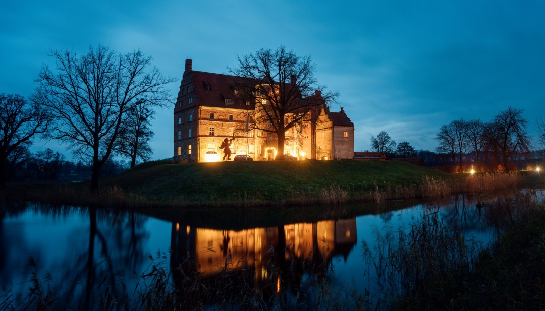 Weihnachtliche Idylle am Schloss Ulrichshusen – festliche Beleuchtung spiegelt sich im Wassergraben und sorgt für eine märchenhafte Abendstimmung., © TMV/Petermann Stimmungsvoll beleuchtetes Schloss Ulrichshusen zur Weihnachtszeit, umgeben von Bäumen und Wassergraben mit Spiegelung im Abendlicht.