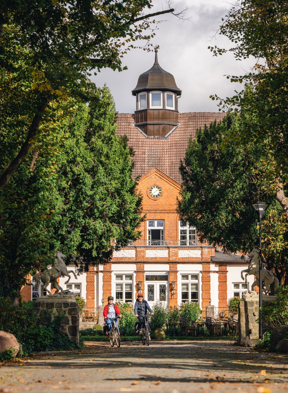 Aktiv unterwegs – Fahrradtour zum beeindruckenden Schloss Basthorst., © TMV/Gross Zwei Radfahrer nähern sich dem Eingang des Schloss Basthorst, eingerahmt von Bäumen und herbstlicher Atmosphäre.