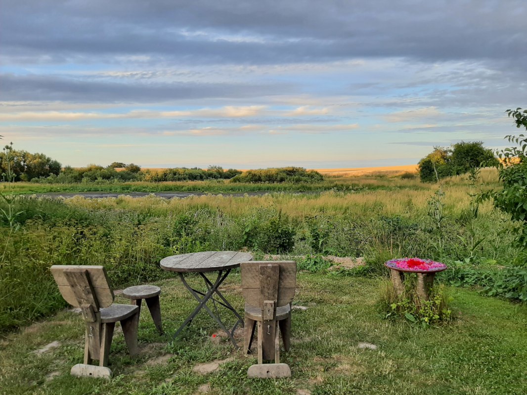Der Sitzplatz am Weiher mit 2 Stühlen, 2 Hockern und 1 Tisch aus Holz. Der Blick geht über den Gartenrand und den zeitweise ausgetrockneten Weiher in die Landschaft. // © Mirabell e.V. Der Sitzplatz am Weiher mit 2 Stühlen, 2 Hockern und 1 Tisch aus Holz. Der Blick geht über den Gartenrand und den zeitweise ausgetrockneten Weiher in die Landschaft. // © Mirabell e.V.