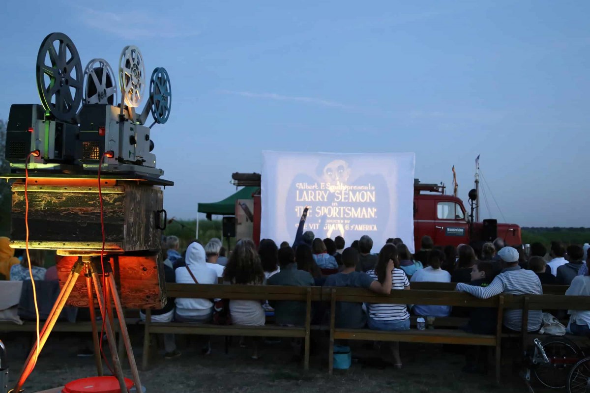 Open-Air-Kinoabend des Wanderkinos Althagen in Ahrenshoop mit einem historischen 16mm- oder 35mm-Filmprojektor im Vordergrund und Publikum vor der Leinwand. // © Tobias Rank Open-Air-Kinoabend des Wanderkinos Althagen in Ahrenshoop mit einem historischen 16mm- oder 35mm-Filmprojektor im Vordergrund und Publikum vor der Leinwand. // © Tobias Rank