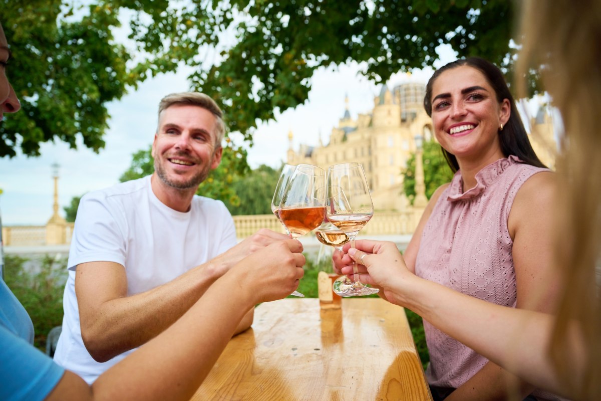 Gasten proosten met een glas wijn tijdens het wijnfeest in Schwerin op de Alte Garten. Op de achtergrond doemt het Slot Schwerin op en vormt zo het bijzondere decor voor het wijnfeest. // &copy; LHSN/Celien Graubaum