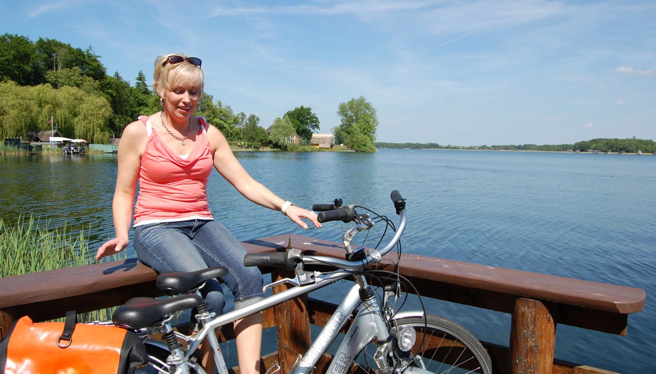 Eine Radfahrerin sitzt auf Geländer am See., © Ralf Tetmeyer, radreisen-mecklenburg Eine Radfahrerin sitzt auf Geländer am See., © Ralf Tetmeyer, radreisen-mecklenburg