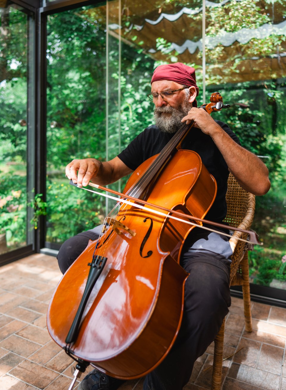 Ein Mann spielt im Wintergarten Cello auf einem Stuhl, konzentriert und ruhig, umgeben von gr&uuml;nem Gartenlicht.