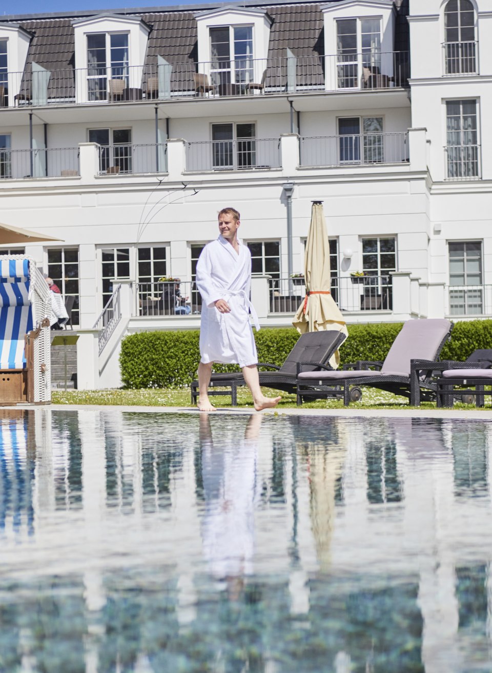 Mann im Bademantel am Pool des Strandhotels Zingst mit Sonnenliegen und Strandkorb vor der Fassade im B&auml;derstil.