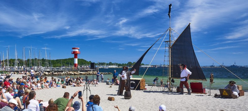 An einem Sandstrand f&uuml;hren mehrere als Piraten verkleidete Personen ein Piratenspektakel vor einem gro&szlig;en Publikum auf. Familien mit Kindern sitzen im Sand und verfolgen die Auff&uuml;hrung, w&auml;hrend im Hintergrund Segelboote, ein Leuchtturm und das Meer zu sehen sind. // &copy; Arthur Niconoculus