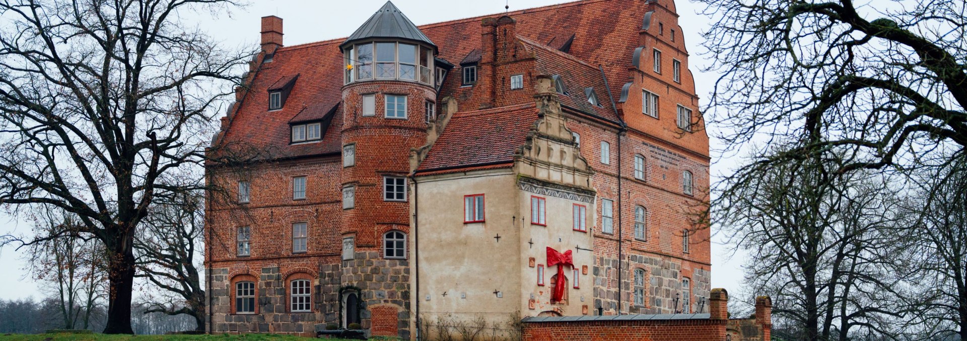 Winterzauber am Schloss Ulrichshusen – Das historische Schloss am Wasser in Mecklenburg erstrahlt festlich geschmückt in der Adventszeit., © TMV/Petermann Außenansicht von Schloss Ulrichshusen in Mecklenburg, mit roter Schleife weihnachtlich dekoriert, umgeben von Bäumen und grüner Winterwiese.