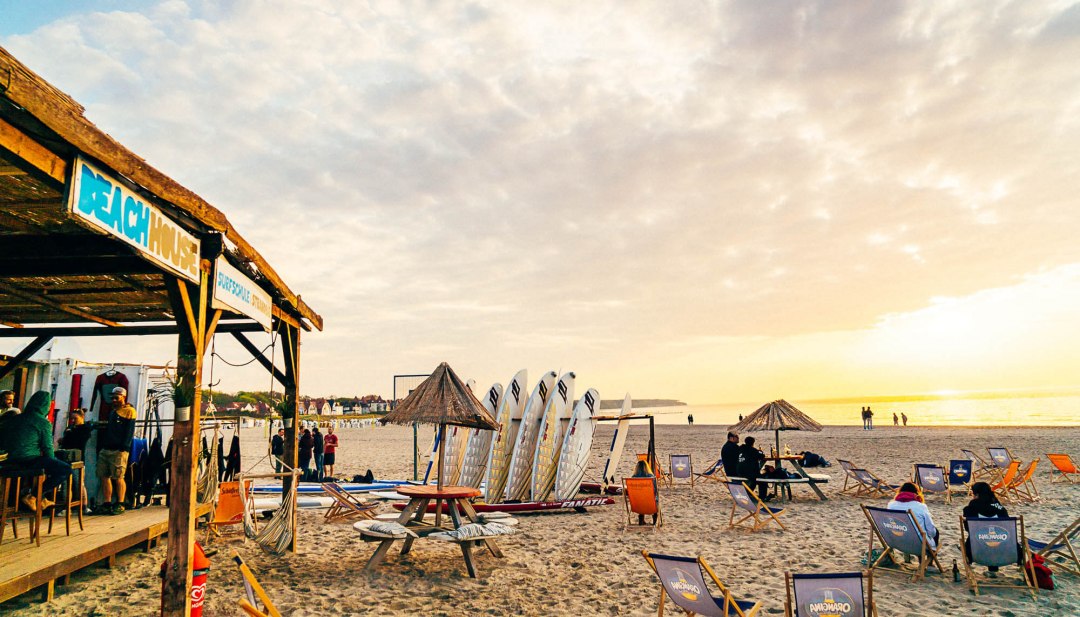 Strandszene mit Beachhouse, Surfbrettern und Sonnenuntergang in Warnemünde, Menschen entspannen in Liegestühlen. // Surfer-Vibes an der Ostsee – Im Supremesurf Beachhouse in Warnemünde genießen Gäste entspannte Stunden direkt am Wasser mit Boards, Bar und Meerblick. // © MV-T/Supremesurf Strandszene mit Beachhouse, Surfbrettern und Sonnenuntergang in Warnemünde, Menschen entspannen in Liegestühlen.