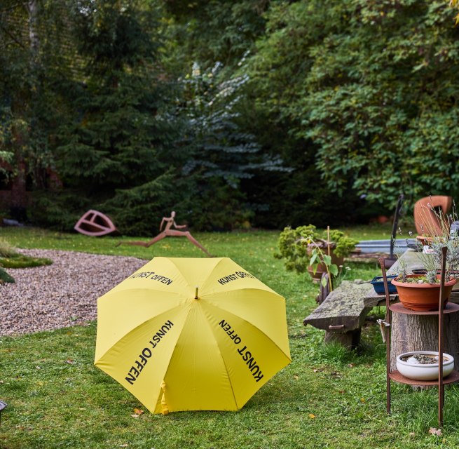 Beeldentuin met gele KunstOffen paraplu en metalen sculpturen bij Kunst-Werk Kummer. Een gele paraplu met de inscriptie "KunstOffen" ligt ervoor; // Bij Kunst-Werk Kummer in Kummer opent Thomas Lehnigk zijn beeldentuin voor KunstOffen. Tussen metalen sculpturen en weelderig groen nodigen houten banken je uit om te vertoeven. Kunst ontmoet natuur - een plek die ruimte biedt voor nieuwe perspectieven met het typische kenmerk - de gele paraplu. // © TVMSN/Borchert Beeldentuin met gele KunstOffen paraplu en metalen sculpturen bij Kunst-Werk Kummer. Een gele paraplu met de inscriptie "KunstOffen" ligt ervoor;