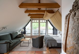 Ein stilvoll eingerichtetes Dachzimmer mit sichtbaren Holzbalken und einem gem&uuml;tlichen Ambiente. Der Raum bietet eine komfortable Sitzecke, ein gro&szlig;es Bett und Zugang zu einem Balkon mit Blick ins Gr&uuml;ne. Warme Terrakotta-B&ouml;den und helle Stoffe runden das gem&uuml;tliche Design ab und laden zum Entspannen ein. // &copy; Hotel Carpe Diem