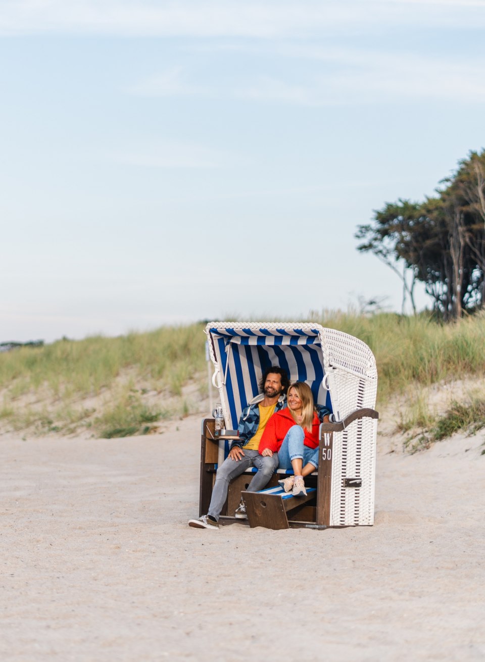 Ein Paar sitzt in einem blau-wei&szlig; gestreiften Strandkorb am Strand von Graal-M&uuml;ritz, umgeben von Sand und D&uuml;nen mit Blick auf die ruhige K&uuml;stenlandschaft.