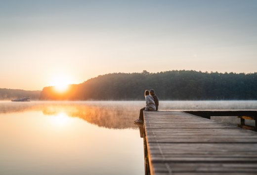Ein Paar sitzt auf einem Holzsteg an einem ruhigen See und schaut in den Sonnenaufgang, umgeben von morgendlichem Nebel.