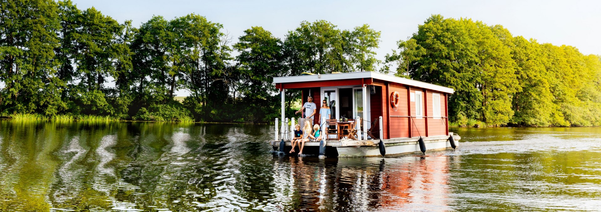 Familie auf einem roten Hausboot auf einem ruhigen See, umgeben von grünen Bäumen und blauem Himmel.