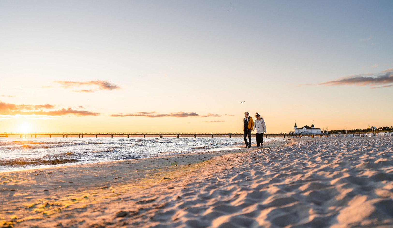 Paar spaziert bei Sonnenuntergang am Strand von Ahlbeck mit Seebr&uuml;cke im Hintergrund.