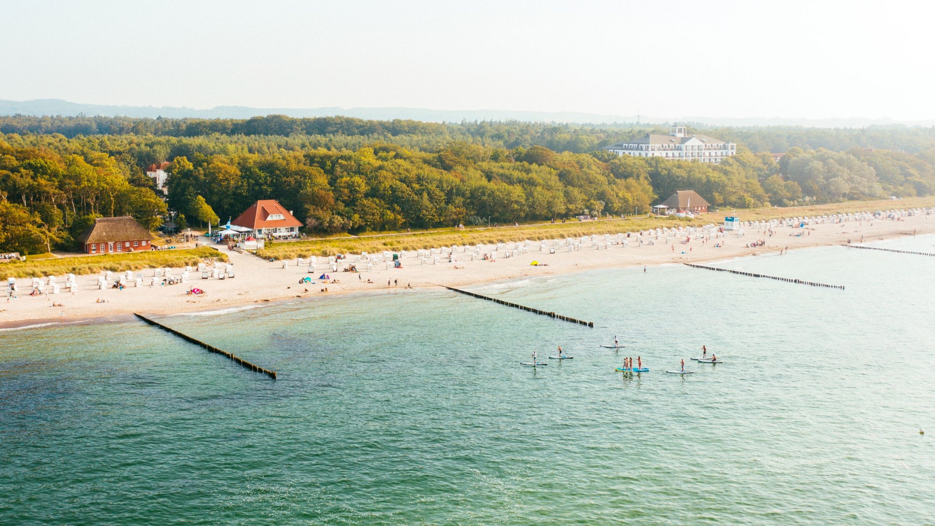 Luchtfoto van het strand in K&uuml;hlungsborn met strandstoelen, badgasten en stand-up paddlers tegen een groene bosrijke achtergrond.