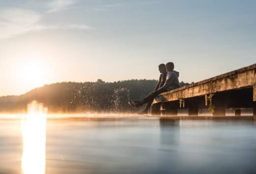 Zwei Personen sitzen im Sonnenaufgang auf einem Steg am Mirower See und lassen die F&uuml;&szlig;e ins glatte Wasser tauchen, w&auml;hrend feiner Nebel &uuml;ber der Oberfl&auml;che schwebt.