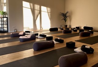 Heller, minimalistischer Yogaraum mit Holzfu&szlig;boden, mehreren Yogamatten, Meditationskissen, Bolstern und Yoga-Bl&ouml;cken. Warmes Sonnenlicht f&auml;llt durch gro&szlig;e Fenster und schafft eine ruhige, entspannende Atmosph&auml;re f&uuml;r Yoga, Meditation und Entspannung. // &copy; MOVA PLACE - Yoga & Co. Studio