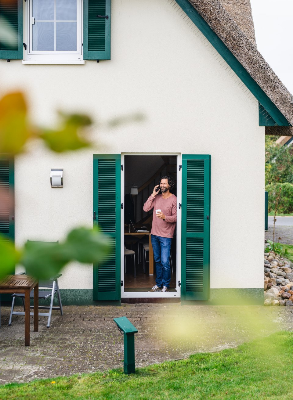 Ein Mann telefoniert und trinkt Kaffee in der T&uuml;r eines Ferienhauses mit gr&uuml;nen Fensterl&auml;den auf der Insel Poel.