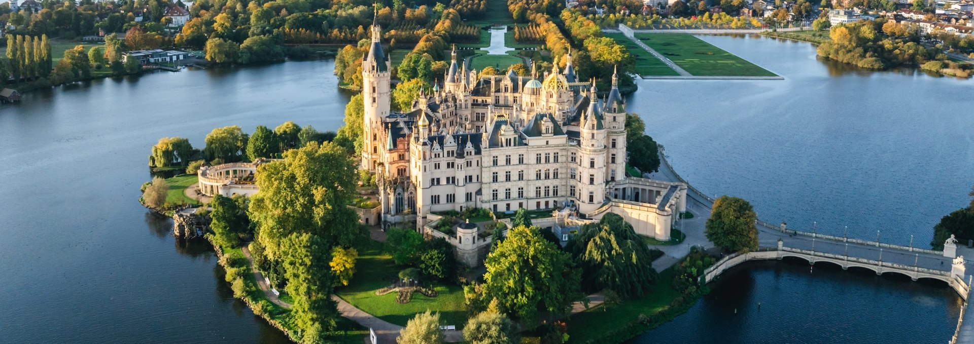Luftaufnahme des Schweriner Schlosses, umgeben von Wasser und herbstlich gefärbten Bäumen im Sonnenaufgang. // Majestätisch und malerisch – Das Schweriner Schloss inmitten der Herbstlandschaft. // © MV-T/Gross Luftaufnahme des Schweriner Schlosses, umgeben von Wasser und herbstlich gefärbten Bäumen im Sonnenaufgang.