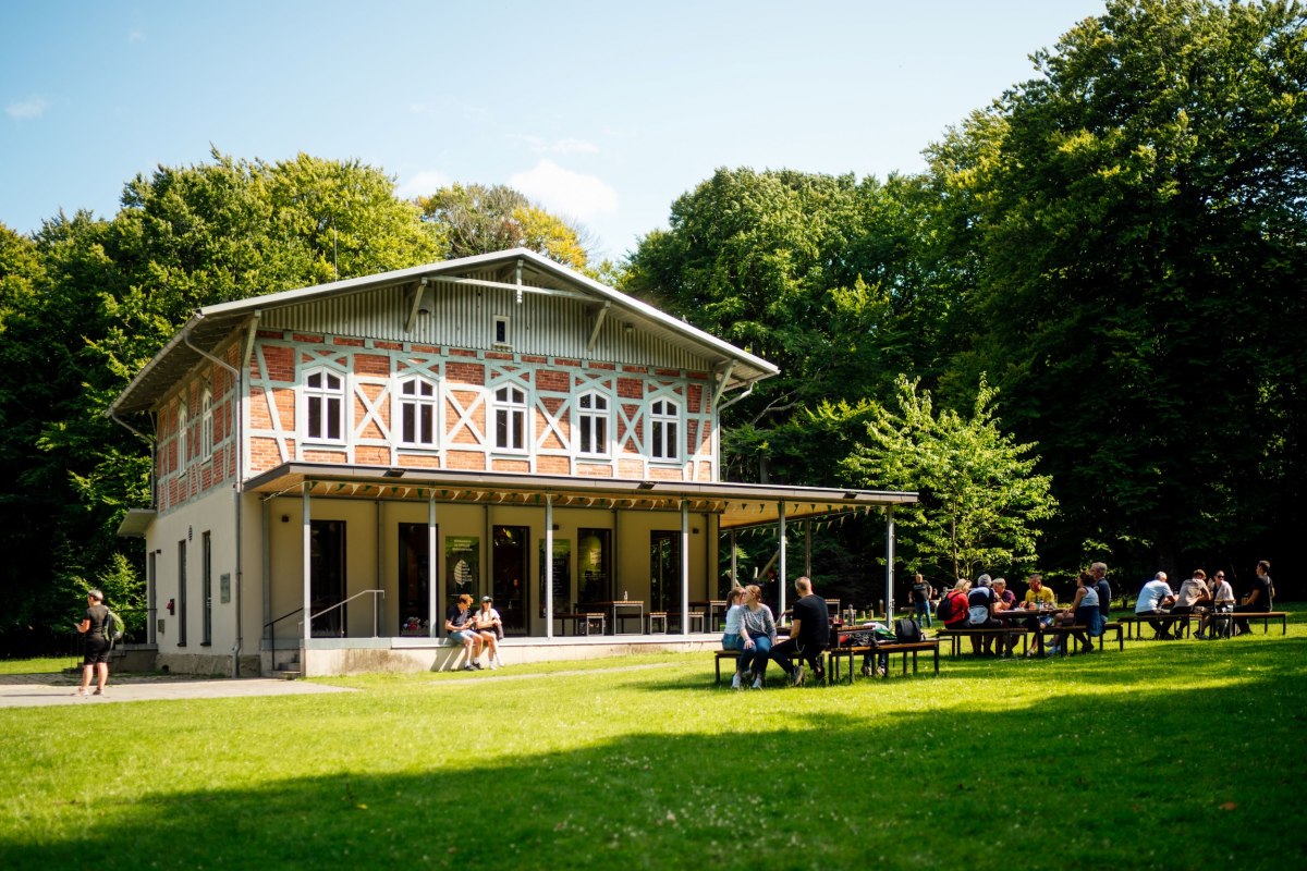 Ein helles Gebäude mit einer Terrasse, vor dem mehrere Personen auf Bänken auf einer grünen Wiese sitzen. // © Nationalpark-Zentrum KÖNIGSSTUHL / Dan Petermann Ein helles Gebäude mit einer Terrasse, vor dem mehrere Personen auf Bänken auf einer grünen Wiese sitzen. // © Nationalpark-Zentrum KÖNIGSSTUHL / Dan Petermann