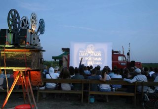 Open-Air-Kinoabend des Wanderkinos Althagen in Ahrenshoop mit einem historischen 16mm- oder 35mm-Filmprojektor im Vordergrund und Publikum vor der Leinwand. // © Tobias Rank Open-Air-Kinoabend des Wanderkinos Althagen in Ahrenshoop mit einem historischen 16mm- oder 35mm-Filmprojektor im Vordergrund und Publikum vor der Leinwand. // © Tobias Rank