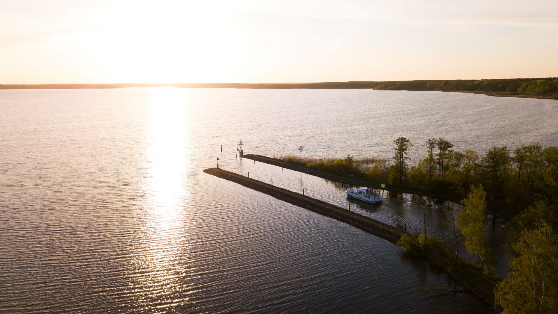 Luftaufnahme des K&ouml;lpinsees bei Sonnenuntergang mit einem Hausboot, das durch einen schmalen Kanal f&auml;hrt, umgeben von B&auml;umen und ruhigem Wasser.