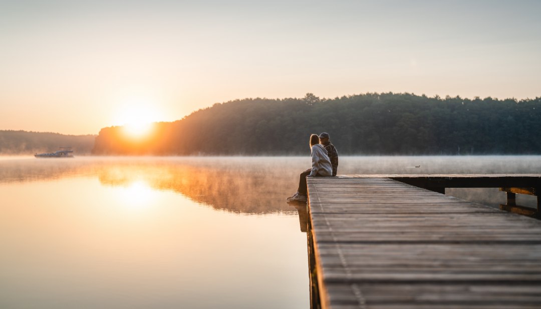 Ein Paar sitzt auf einem Holzsteg an einem ruhigen See und schaut in den Sonnenaufgang, umgeben von morgendlichem Nebel. // Magische Morgenstimmung – Ein Paar genießt die Ruhe und den Sonnenaufgang auf einem Steg am stillen See, während sich Nebel über das Wasser legt. // © MV-T/Gross Ein Paar sitzt auf einem Holzsteg an einem ruhigen See und schaut in den Sonnenaufgang, umgeben von morgendlichem Nebel.