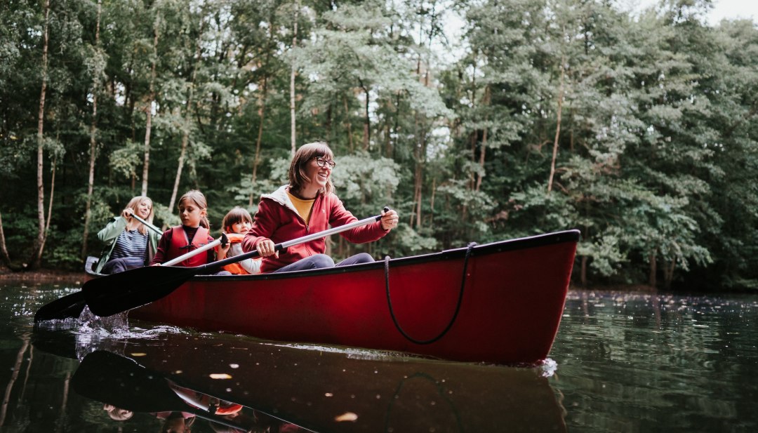 Een lachende vrouw peddelt in een rode kano; achter haar zitten twee kinderen met zwemvesten op het rustige water. // Gekabbel, stilte, bosgroen: in het Nationaal Park Müritz glijden gezinnen in een kano over het stille water. // © 1000seen Een lachende vrouw peddelt in een rode kano; achter haar zitten twee kinderen met zwemvesten op het rustige water.
