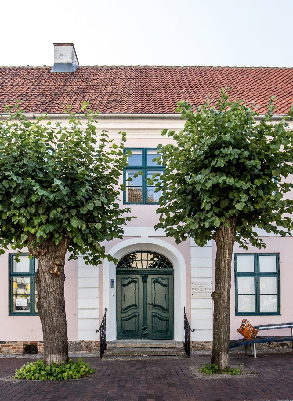 Ein rosa Gebäude mit grünen Fenstern steht hinter zwei belaubten Linden, am Haus hängt ein Schild mit der Aufschrift Rungehaus. // Vor der rosa Fassade des Rungehauses in Wolgast stehen zwei Linden. Eine Gedenktafel neben der Tür erinnert an den Maler Philipp Otto Runge. // © MV-T/Krauss Ein rosa Gebäude mit grünen Fenstern steht hinter zwei belaubten Linden, am Haus hängt ein Schild mit der Aufschrift Rungehaus.