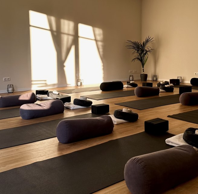 Heller, minimalistischer Yogaraum mit Holzfu&szlig;boden, mehreren Yogamatten, Meditationskissen, Bolstern und Yoga-Bl&ouml;cken. Warmes Sonnenlicht f&auml;llt durch gro&szlig;e Fenster und schafft eine ruhige, entspannende Atmosph&auml;re f&uuml;r Yoga, Meditation und Entspannung. // &copy; MOVA PLACE - Yoga & Co. Studio