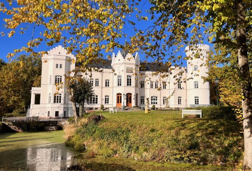 Vor Schloss Schlemmin leuchten Herbstblätter über dem Wassergraben, die weiße Fassade steht vor blauem Himmel. // Weiße Schlossfassade, goldenes Laub, stiller Wassergraben – Schloss Schlemmin zeigt Mecklenburg-Vorpommern von seiner klaren Seite. // © Schloss Schlemmin Vor Schloss Schlemmin leuchten Herbstblätter über dem Wassergraben, die weiße Fassade steht vor blauem Himmel.