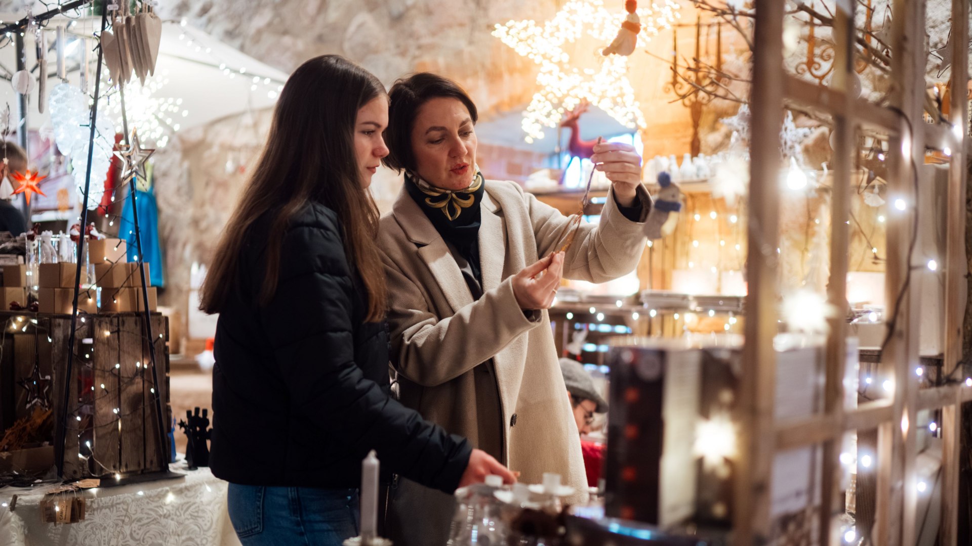 Zwei Frauen auf einem festlich geschmückten Weihnachtsmarkt betrachten liebevoll gestaltete Dekorationen, umgeben von Lichterglanz und winterlicher Atmosphäre. // Festliche Magie erleben handgefertigten Weihnachtsschmuck in stimmungsvoller Beleuchtung bewundern // © MV-T/Petermann Zwei Frauen auf einem festlich geschmückten Weihnachtsmarkt betrachten liebevoll gestaltete Dekorationen, umgeben von Lichterglanz und winterlicher Atmosphäre.