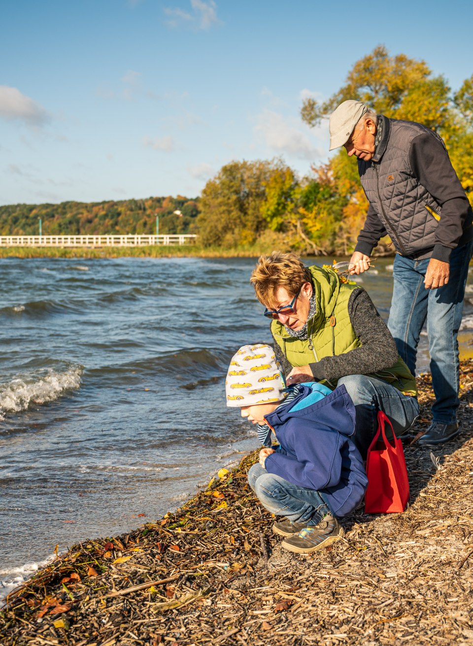Gro&szlig;eltern hocken mit Enkelsohn am Wasser des Tollensesees im Herbst