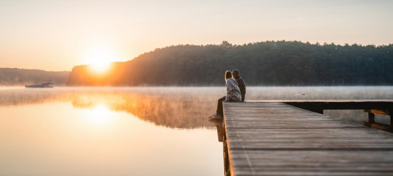 Ein Paar sitzt auf einem Steg zum Sonnenuntergang am Mirower See. Im Hintergrund f&auml;hrt ein Hausboot vorbei.