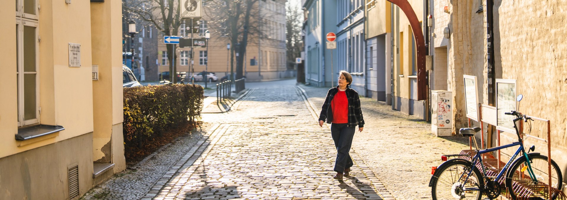 Frau spaziert durch kopfsteingepflasterte Altstadtgasse in Greifswald bei Sonnenschein.