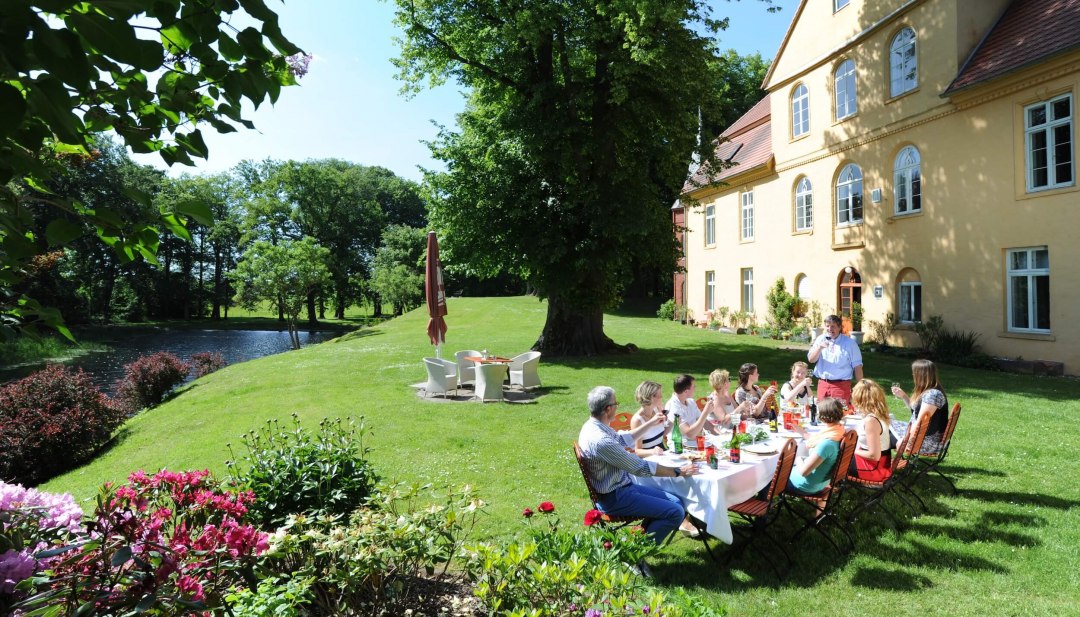 Personen sitzen in einem Garten vor einem Gutshaus mit einer langen Tafel und picknicken bei Sonnenschein.
