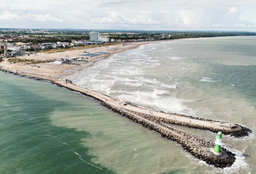 Luftaufnahme vom Strand Warnemünde mit Mole, Leuchtturm und Ostsee bei sanftem Wellengang. // Die Ostsee legt sich in sanften Wellen an den breiten Sandstrand von Warnemünde. Die steinerne Mole führt hinaus aufs Meer, gekrönt vom grün-weißen Leuchtturm. Strandkörbe reihen sich entlang der Küste, während sich dahinter die Silhouette des Seebads mit historischen Gebäuden und modernen Hotels abzeichnet. // © MV-T/Scholz-Witzel Luftaufnahme vom Strand Warnemünde mit Mole, Leuchtturm und Ostsee bei sanftem Wellengang.