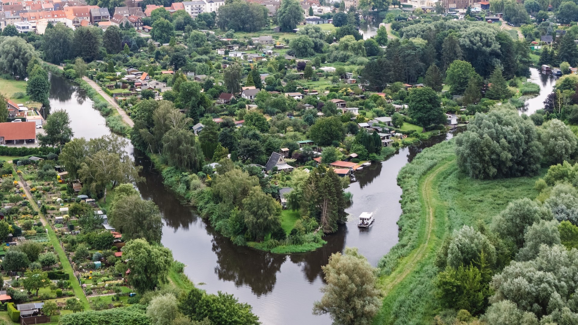Eine Luftaufnahme zeigt die kurvige Elde mit einem Hausboot, umgeben von gr&uuml;nen G&auml;rten und der Stadt Parchim im Hintergrund.