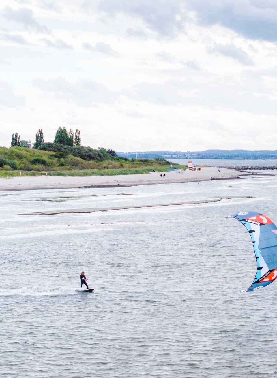 Ein Kitesurfer gleitet vor der K&uuml;ste von Timmendorf auf Poel &uuml;ber das Wasser, im Hintergrund eine gr&uuml;ne D&uuml;nenlandschaft.
