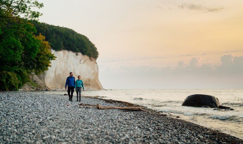 Ein Paar spaziert am Fu&szlig;e der ber&uuml;hmten Kreidefelsen auf der Insel R&uuml;gen entlang, w&auml;hrend der Sonnenuntergang den Himmel in sanften Farben erstrahlen l&auml;sst.