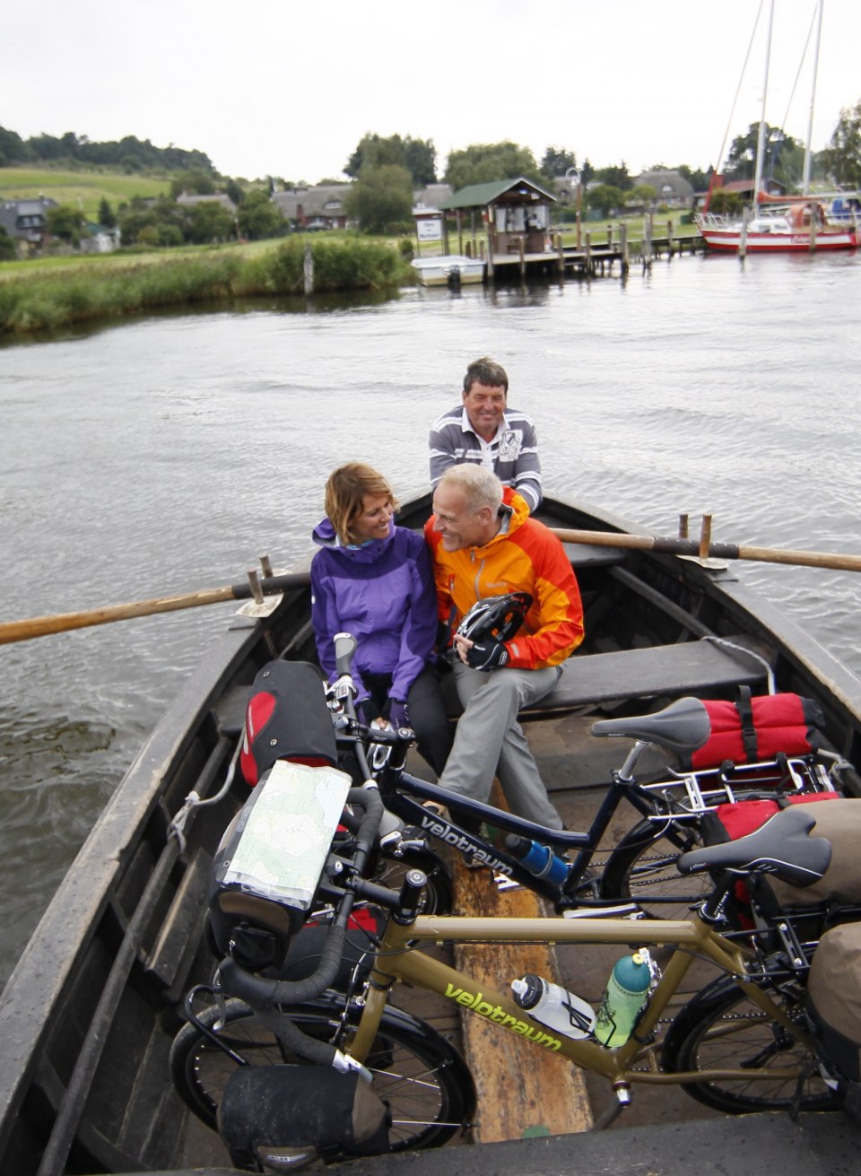Drei Personen mit Fahrrädern auf einer Ruderfähre am Baaber Bollwerk, umgeben von Wasser, mit Blick auf grüne Ufer und einen kleinen Hafen im Hintergrund. // Eine idyllische Überfahrt: Radfahrer nutzen die traditionelle Ruderfähre am Baaber Bollwerk auf Rügen, um den Bodden zu überqueren und die Schönheit der Natur zu genießen. // © MV-T/outdoor-visions.com Drei Personen mit Fahrrädern auf einer Ruderfähre am Baaber Bollwerk, umgeben von Wasser, mit Blick auf grüne Ufer und einen kleinen Hafen im Hintergrund.