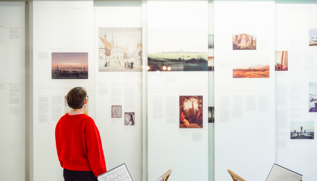 Besucherin betrachtet Caspar David Friedrich Reproduktionen im Ausstellungsraum in Greifswald. // Im Caspar David Friedrich Zentrum in Greifswald wird die Welt des Romantikers greifbar. Reproduktionen seiner Meisterwerke, biografische Einblicke und interaktive Stationen laden zum Verweilen ein. Hier lässt sich nachspüren, wie der Künstler seine Heimat Vorpommern in zeitlose Kunst verwandelte. // © MV-T/Gross Besucherin betrachtet Caspar David Friedrich Reproduktionen im Ausstellungsraum in Greifswald.