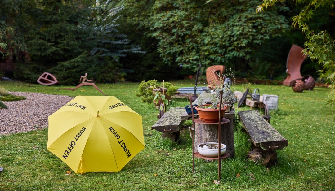 Skulpturengarten mit gelbem KunstOffen-Schirm und Metallskulpturen im Kunst-Werk Kummer. Davor liegt ein gelber Regenschirm mit der Aufschrift "KunstOffen"