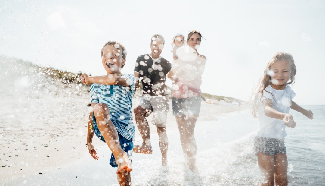 Eine Familie l&auml;uft lachend durch die flachen Ostseewellen am Strand von Dierhagen, w&auml;hrend Wasser in der Sonne lebendig aufspritzt.