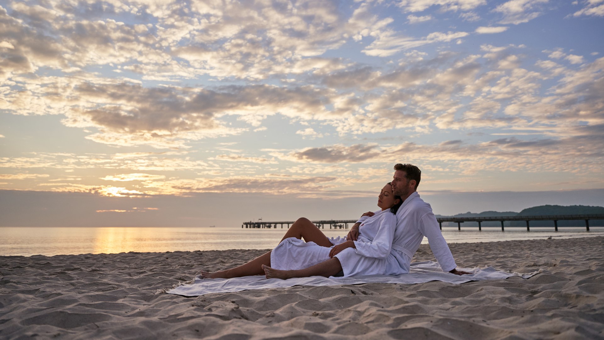 Zwei Personen sitzen am Strand im bademantel mit der Seebrücke Binz im Hintergrund // Ostseefeeling am Strand im Bademantel // © Kurhaus Binz Zwei Personen sitzen am Strand im bademantel mit der Seebrücke Binz im Hintergrund