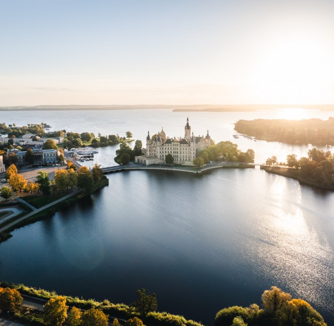 Schweriner Schloss auf Insel im See bei Sonnenaufgang, umgeben von B&auml;umen und Wasserfl&auml;chen.
