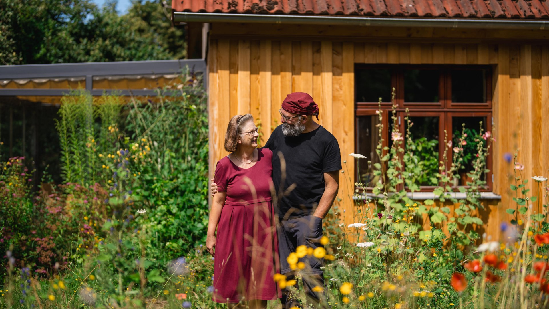 Ein Paar steht in einem bl&uuml;henden Garten vor einem Holzhaus und l&auml;chelt sich an, bunte Wildblumen im Vordergrund.