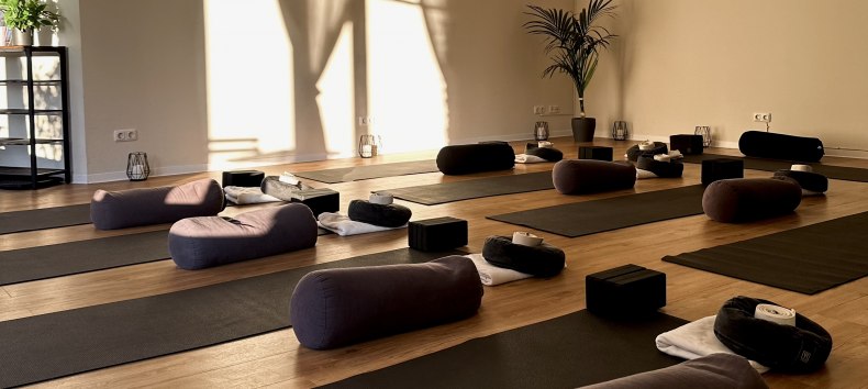 Heller, minimalistischer Yogaraum mit Holzfu&szlig;boden, mehreren Yogamatten, Meditationskissen, Bolstern und Yoga-Bl&ouml;cken. Warmes Sonnenlicht f&auml;llt durch gro&szlig;e Fenster und schafft eine ruhige, entspannende Atmosph&auml;re f&uuml;r Yoga, Meditation und Entspannung. // &copy; MOVA PLACE - Yoga & Co. Studio