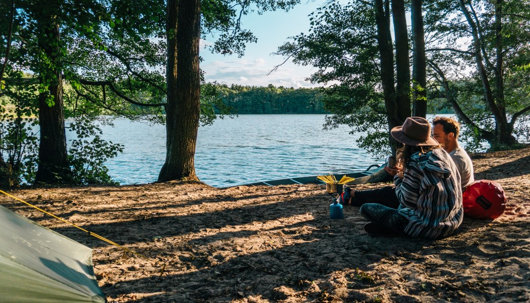 Ein Paar sitzt am Wasser mit einem Gaskocher und kocht Nudeln am Campingplatz Hexenwäldchen