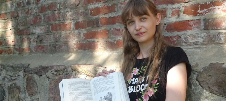 Jose Saefkow sitzt im schwarzen Shirt vor einer alten Klinkerwand und zeigt ihr Buch // &copy; Josi Saefkow